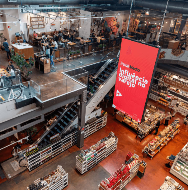 Fotografia de um grande comércio, com vários produtos e consumidores. Uma tv exibe Retail Media - Influência digital no varejo